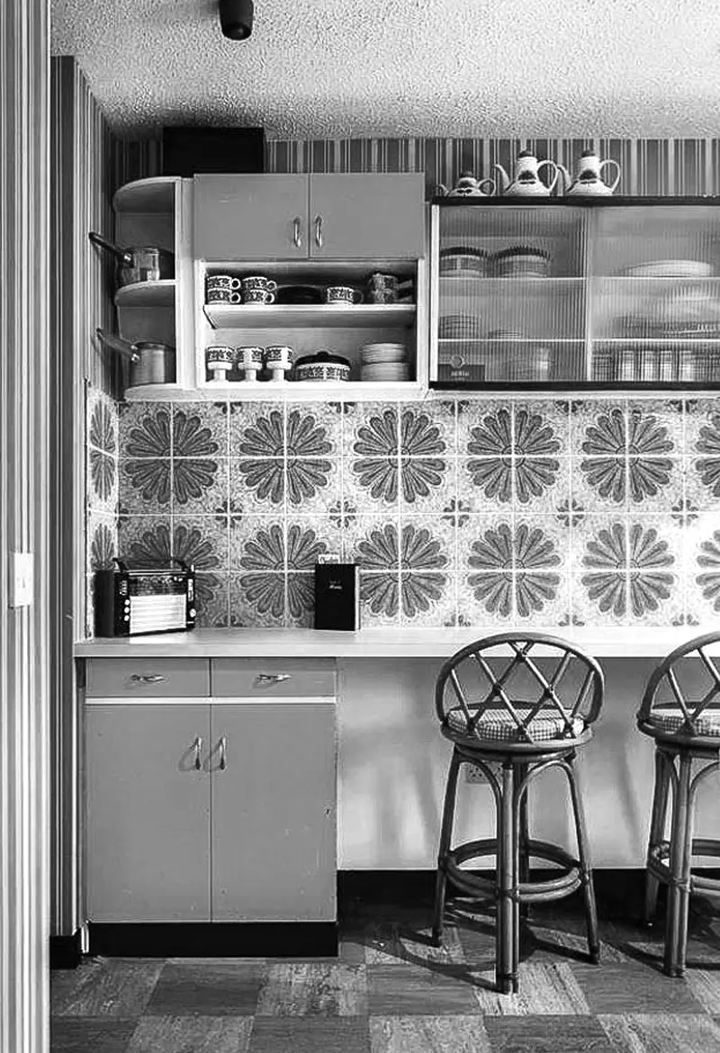 cocina de los años 70 con azulejos con dibujo de flor de manzanilla armarios colgados para menaje de te y encimera blanco bancos para sentarse echos con madera de mimbre